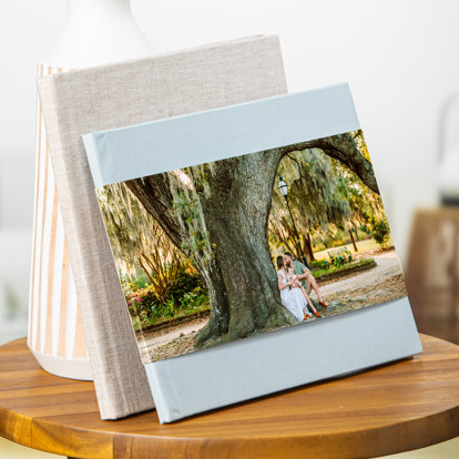 Two photo books leaning up against a lamp on a table. The one in front is a hardcover photo book with light blue linen cover and skinny dustjacket featuring a picture of a family sitting under a large tree in the park.