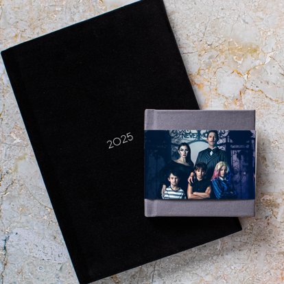 A closed photo book with a dark linen cover and skinny dust jacket stacked on top of a closed photo album with a black velvet cover and foil stamped debossed text reading "2025."