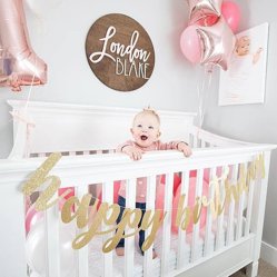 A baby standing in a crib under a round name sign, first-birthday balloons behind