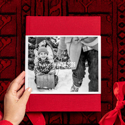 A closed hardcover photo book with a red linen cover and a skinny dustjacket featuring a photo of kids being pulled on a toboggan in the snow. 