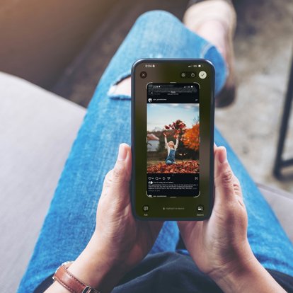 Hands holding a phone so you can see the screen. The screen shows a screenshot off social media featuring a little girl playing in a pile of leaves.