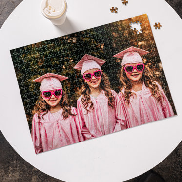 A custom jigsaw puzzle from Mpix sitting on a table featuring a photo of three children in graduation caps and gowns.