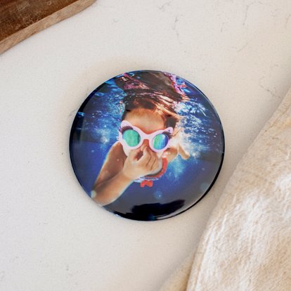 A button with a personalized photo of a girl swimming underwater with bright blue goggles and a protective coating