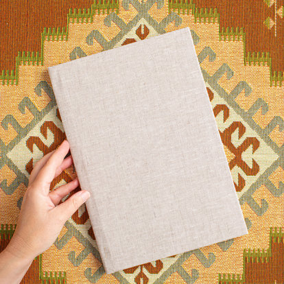 A closed 8.5x11 photo book with a sand color linen cover lying on a colorful tablecloth. 