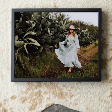 A framed print from Mpix with a thin black metal frame featuring a girl walking through a field wearing a blue flowing dress and a sun hat for her graduation photos. 