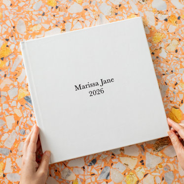 Hands holding a Signature Photo Album from Mpix with a polar white leather cover and black debossing that reads "Marissa Jane 2026"