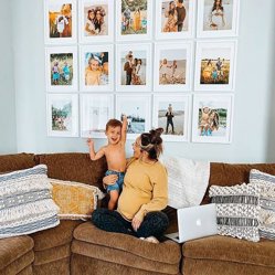 A mother and toddler on a couch below a large grid of framed photos