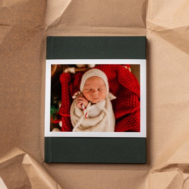 A personalized hardcover photo book with a green linen cover and skinny dust jacket with a personalized photo of a baby resting in a Christmas setting resting on kraft paper. 