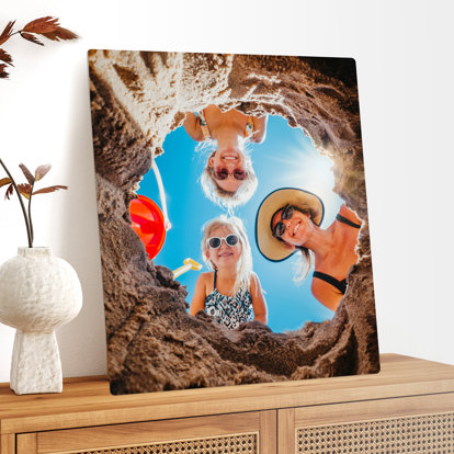 A large metal print displayed on a side table featuring a photo of three smiling people looking down into a sand hole at the beach. The photo is taken from inside the hole looking up. 