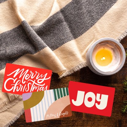 Three Christmas themed Mpix gift cards sitting on a table next to a candle and a blanket. 