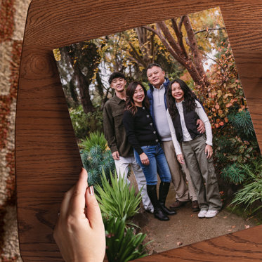 An 8x10 giclee photo print set on a dark wood table featuring a family of four standing on a walkway surrounded by lush green plants and trees.