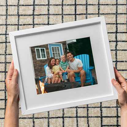 A framed print featuring a couple and their two young children sitting on blue adirondack chairs by a firepit. The photo has a white window mat and white metal frame.