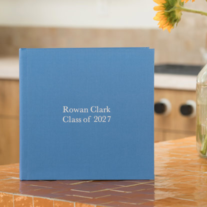 A closed custom fabric photo book with a bright blue cover and the title ‘Rowen Clark Class of 2027’ in silver debossing.
