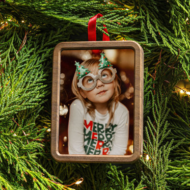 A framed photo ornament hanging on a Christmas tree. The photo is of a little girl in a 'Merry' shirt wearing circular glasses with Christmas trees on top.
