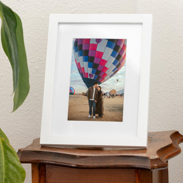 A tabletop 5x7 framed photo with white mat and white frame featuring a couple standing in front of a hot air balloon.