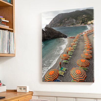 A large canvas print hanging on the wall featuring a shoreline at the edge of a mountain lined with orange and green beach umbrellas.