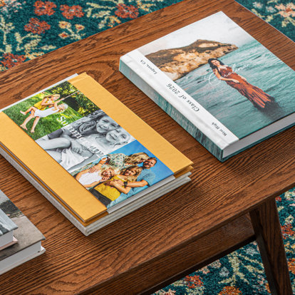 A table with three different small stacks of photo books. At the top of one stack is a graduation photo book with a custom cover. At the top of the second stack is a hardcover photo book with a caramel color linen cover and dustjacket featuring a family's summer photos.