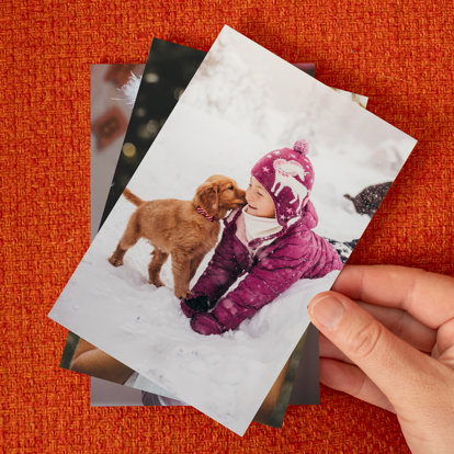Hands holding a 4x6 photo print of a toddler girl playing in the snow with her golden retriever puppy.