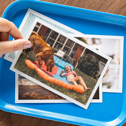 A few photo prints scattered on a small blue tray. The top photo print is of a little girl playing on a slip-n-side with her golden retriever.