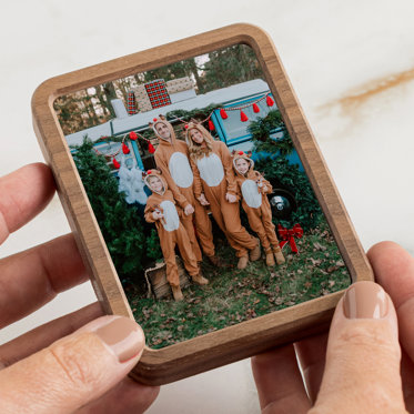 Hands holding a rectangular shaped framed photo magnet of a family all dressed in reindeer onesies in front of a Christmas tree farm setting.