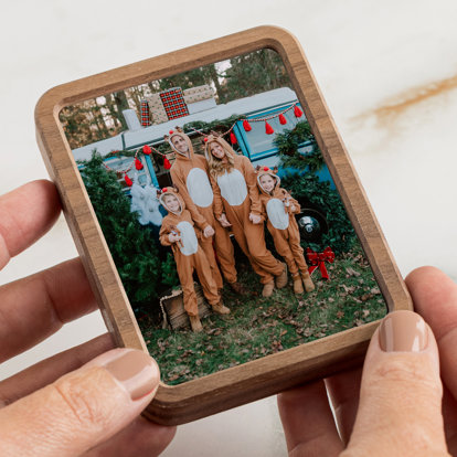 Hands holding a rectangular shaped framed photo magnet of a family all dressed in reindeer onesies in front of a Christmas tree farm setting.