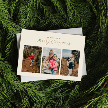 A Photo Christmas Card laying on an envelope and a green wreath. The Card has colorful text at the top reading "We Wish You A Merry Christmas", with three photos below of a family in a Christmas tree field.