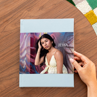 A closed hardcover photo book with a light blue linen cover and a skinny dust jacket featuring a photo of a high school graduate and her name 'Jessa' in text. 