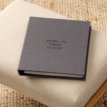 A closed photo album with pockets showing the dark graphite gray linen cover and debossed text in black foil that reads "Janelle + Eli Patrick 1/1/2023"