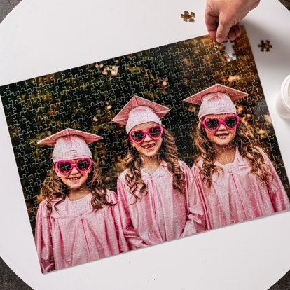 An assembled photo puzzle featuring three young girls in pink graduation gowns and caps with pink sunglasses on.