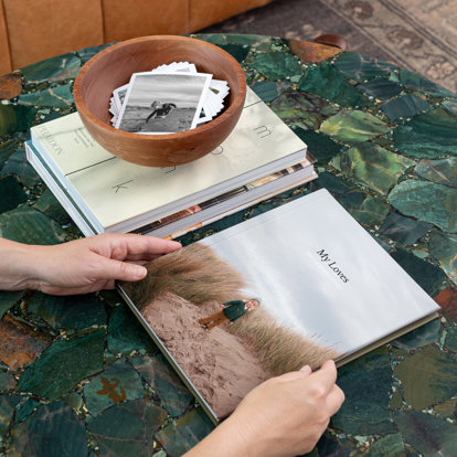 Hands holding a closed photo book with a custom cover on a table next to two other closed photo books with a wooden bowl on top of them that has a stack of photo prints inside.