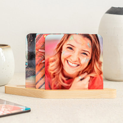 A set of four personalized coasters in a display stand. The front coaster features a woman smiling with streaks of bright colored chalk on her face. 