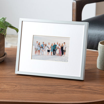 A small framed print featuring a large family standing on a white sandy beach. The frame is a thin, flat silver metal with a white window mat.
