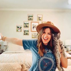 A woman in a hat holding a cat in front of a wall of small framed prints