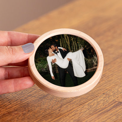 A hand holding a circular framed magnet featuring a photo of a groom holding his bride in his arms and kissing her.