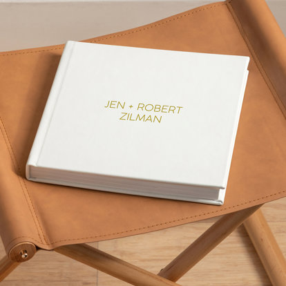 A closed wedding photo album with a white leather cover and the title 'Jen + Robert Zilman' in gold debossing on the front cover.