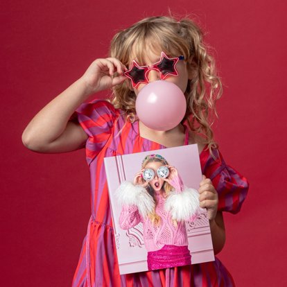 A young girl wearing star-shaped sunglasses and with a bubble gum bubble holding a photo print.