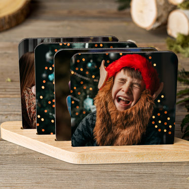 Set of four photo coasters from Mpix in wooden display stand featuring different holiday images, the first one showing is of a kid in a fuzzy santa hat with a big beard and pointy ears. 