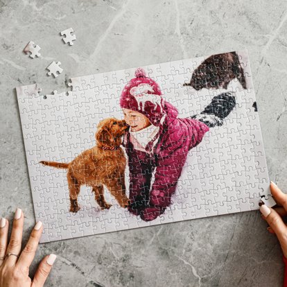 Hands assembling a custom jigsaw photo puzzle that is personalized with an image of a young child playing with a puppy dog in the snow.