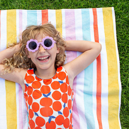 A young girl wearing flower-shaped sunglasses lying on a colorful striped personalized beach towel outside in the grass. 