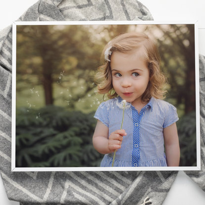 a framed photo print of a young girl in the wood blowing a dandelion professionally framed with a thin silver metal frame. 