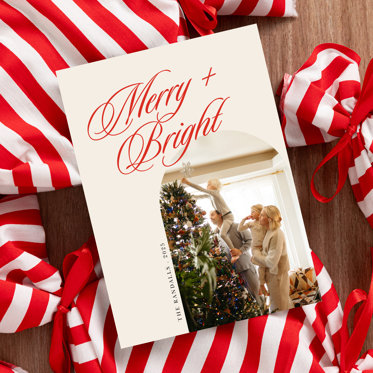 A Christmas Photo Card from Mpix with a photo of a family decorating a Christmas tree, personalized text, and the greeting "Merry + Bright" in large red script font at the top.