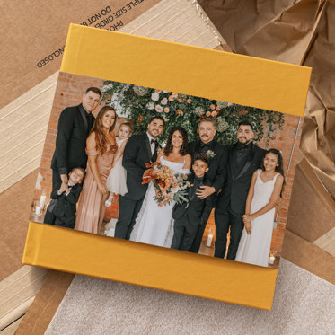 A wedding photo book with a caramel color linen cover and skinny dust jacket featuring a photo of a bride and groom with their family on their wedding day. 