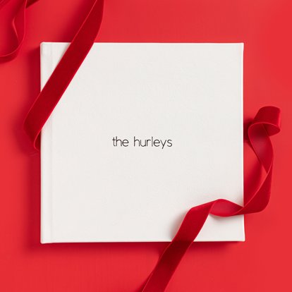 A closed photo album with a polar white leather cover and black foil stamped text that reads "the hurleys."