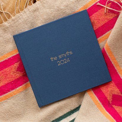 A closed photo book with a windsor blue linen cover and gold foil debossed text that reads "they smyths 2024".