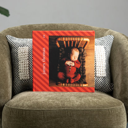 A closed hardcover photo book with a custom cover featuring a diagonal red stripe background and a photo of a newborn baby sleeping in a tiny bed. 