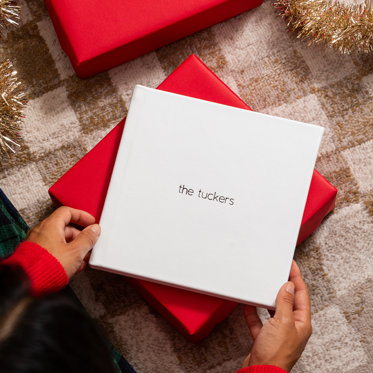 Hands holding a closed photo album from Mpix with a polar white leather cover and black foil debossed text that reads "the tuckers" 