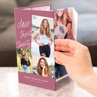 A hand opening an accordion photo book that is standing up on a table and featuring images of a high school senior girl.