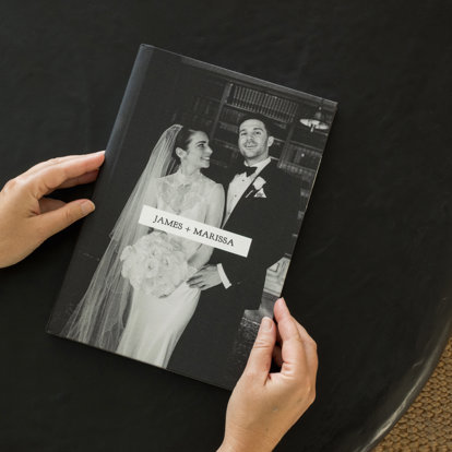 A closed 8.5x11" custom fabric photo book featuring a black & white photo of a bride and groom. The title reads 'James + Marissa'.