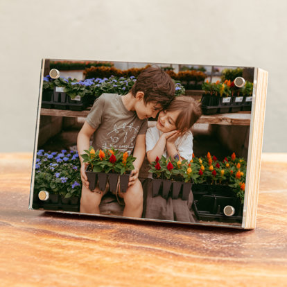 A birch photo block displayed on a table featuring a photo of a brother and sister sitting next to each other at a nursery with a tray of flowers on their laps.