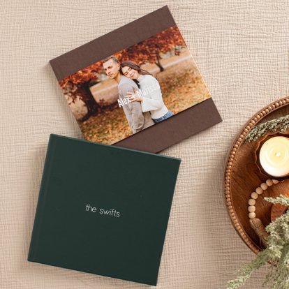 A closed photo album with a dark green leather cover and personalized foil stamped text and a closed photo book with a dark brown linen cover with a photo dust jacket.